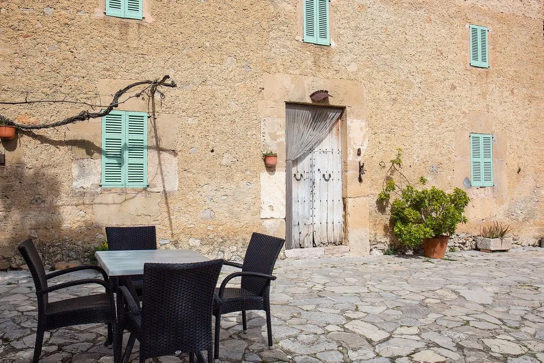 A shaded patio with a table and chairs against the weathered facade, a painted door and potted plants