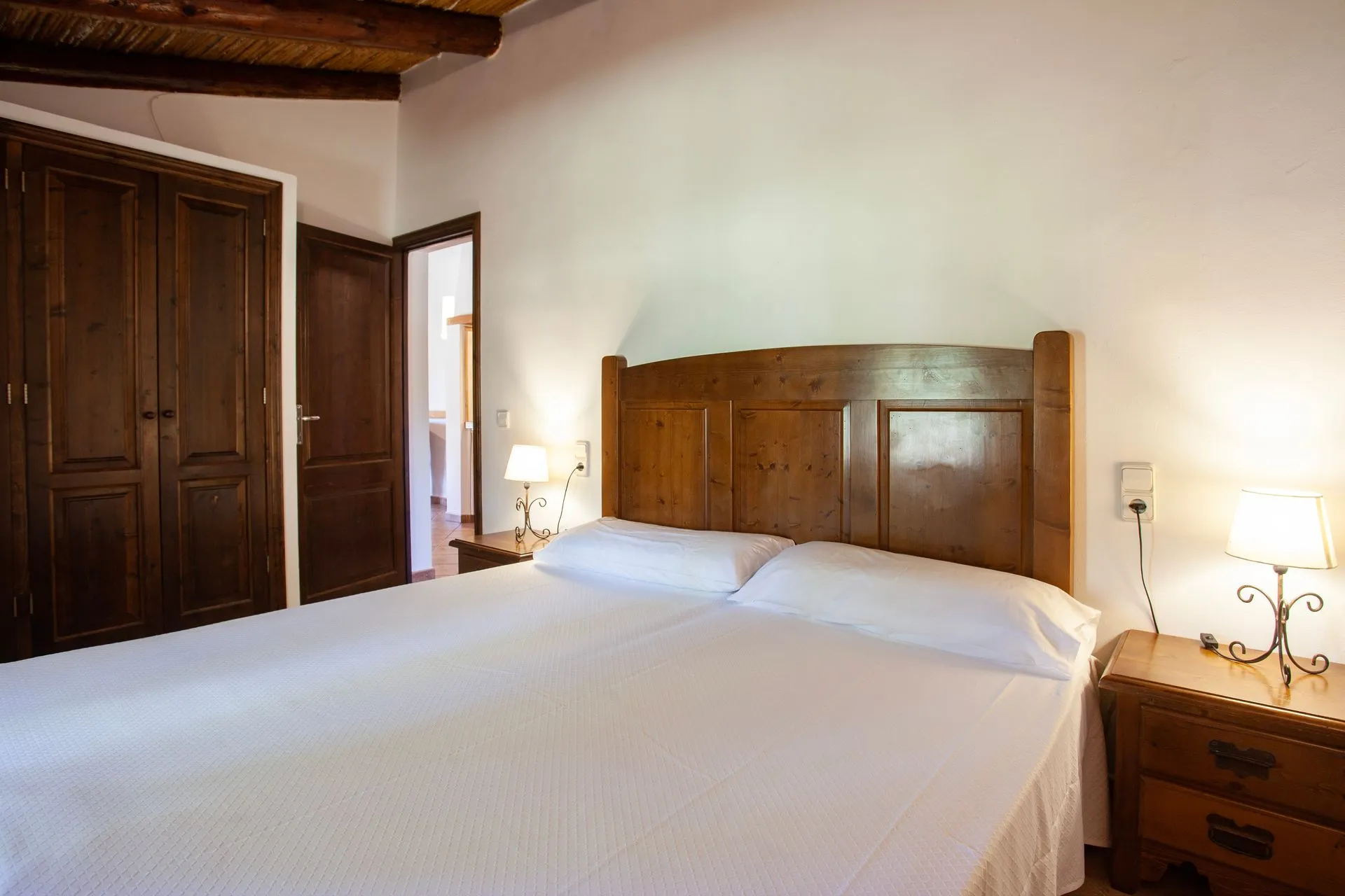 A bedroom with a wide bed, a carved wooden headboard and a beamed ceiling